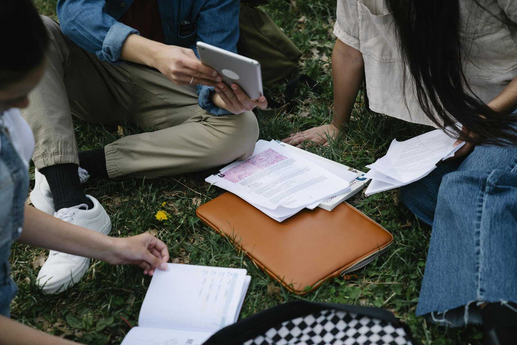 A group of students studying together outside, using a tablet and papers on the grass.