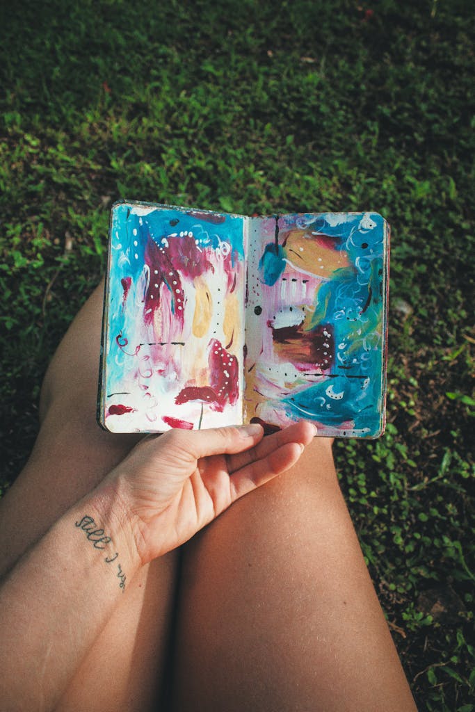A person holding an abstractly painted sketchbook while sitting on grass, demonstrating creativity.
