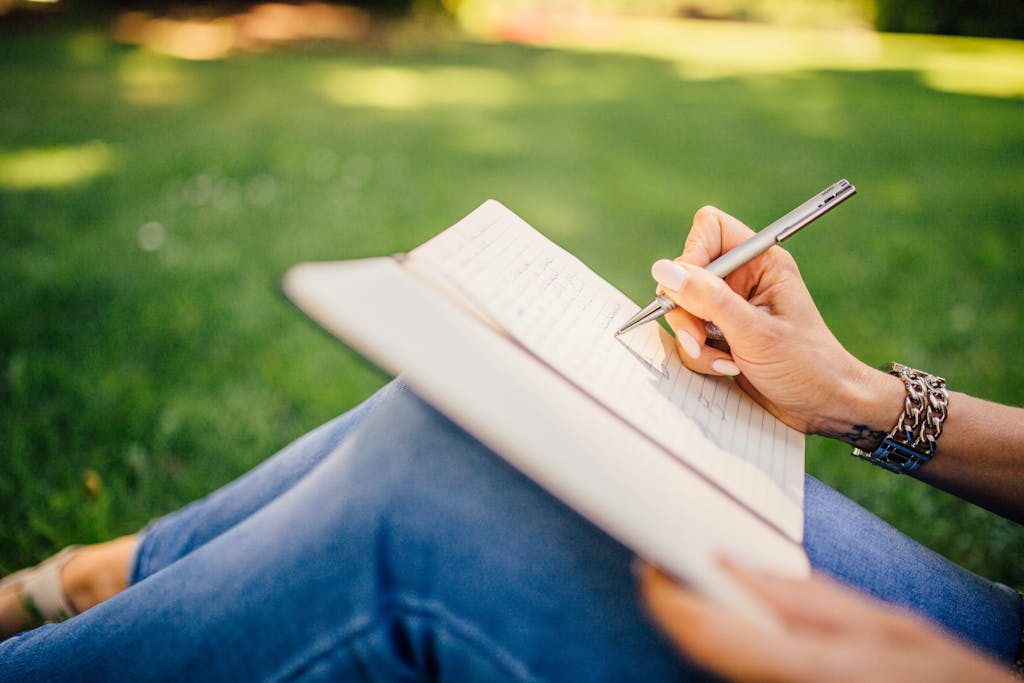 Journaling for overwhelm, a person writes in a notebook on a sunny day. Focus on hands and pen outdoors.