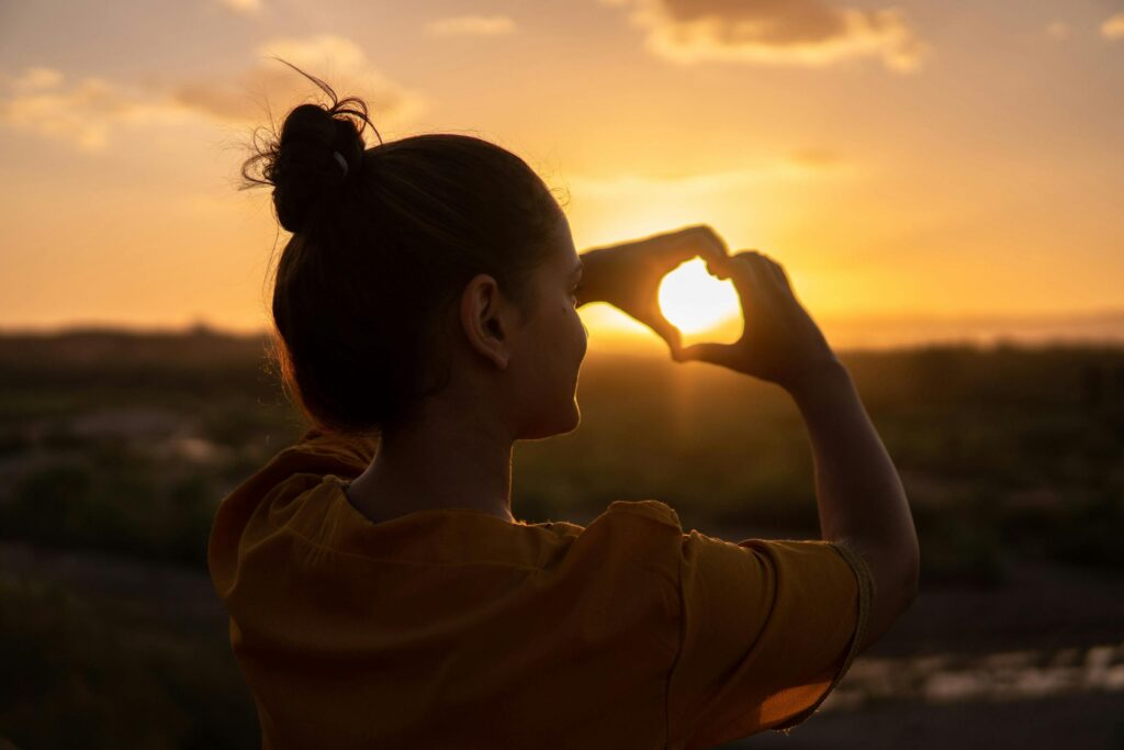 Journaling practices - A silhouette of a young woman forming a heart shape with her hands at sunset.